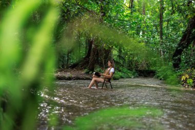 Asyalı bir kadın derede bir sandalyede oturmuş elinde bir kitapla kahve içiyor ve tatil konseptinde rahatlıyor..