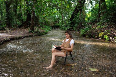 Asyalı bir kadın derede bir sandalyede oturmuş elinde bir kitapla kahve içiyor ve tatil konseptinde rahatlıyor..