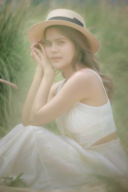 Beyaz elbiseli ve şapkalı Asyalı güzel bir kadın portresi çayırda oturuyor, çiçek yeşili çimlerde dinleniyor, eski moda ve eski moda. yumuşak odak ve sanat konsepti duygusu.