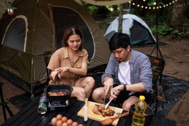 Genç Asyalı çift akşam vakti doğa parkında kamp yapmak için yemek hazırlıyor. Seyahat, macera ve tatil konsepti