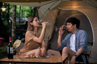 Genç Asyalı çift, doğanın ortasında kamp alanında şarap içip içki içiyorlar.