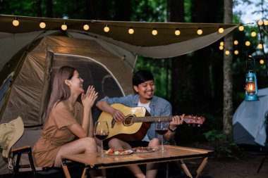 Genç Asyalı çift gitar çalarken gülüyor ve kamp yemeğinin tadını çıkarıyor.