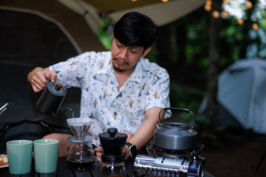 Kamplarındaki maceraperest genç Asyalı adam sabah kahvaltısı için doğal parkta kahve hazırlıyor..
