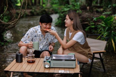 Genç bir çift iskemlede oturuyor ve derede dinleniyor. Sabahları kahve içip kadeh kaldırıyor. Kamp yapıyor, tatil konseptinde dinleniyor..