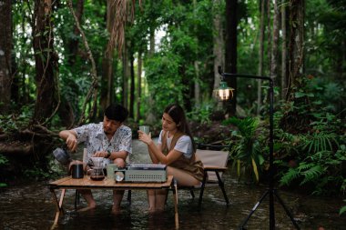 Genç bir çift iskemlede oturuyor ve derede dinleniyor. Sabahları kahve içip kadeh kaldırıyor. Kamp yapıyor, tatil konseptinde dinleniyor..