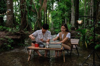 Genç bir çift iskemlede oturuyor ve derede dinleniyor. Sabahları kahve içip kadeh kaldırıyor. Kamp yapıyor, tatil konseptinde dinleniyor..