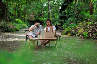 Genç Asyalı çift, tatilde, serbest ve rahatlatıcı bir konseptte, tropikal bir ormanın ortasında, dizüstü bilgisayar kullanarak ve birlikte kahve içerek tahta sandalyelerde oturmaktan zevk alıyorlar.