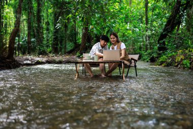 Genç Asyalı çift, tatilde, serbest ve rahatlatıcı bir konseptte, tropikal bir ormanın ortasında, dizüstü bilgisayar kullanarak ve birlikte kahve içerek tahta sandalyelerde oturmaktan zevk alıyorlar.
