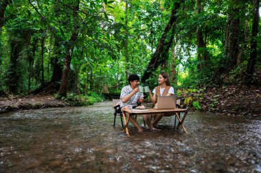Genç Asyalı çift, tatilde, serbest ve rahatlatıcı bir konseptte, tropikal bir ormanın ortasında, dizüstü bilgisayar kullanarak ve birlikte kahve içerek tahta sandalyelerde oturmaktan zevk alıyorlar.