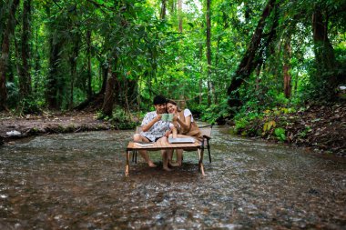 Rahatla ve romantik zaman kavramı, genç Asyalı çift tatilde tropikal ormandaki bir derenin ortasında birlikte oturup kahve içmekten zevk alıyorlar..
