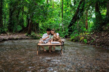 Rahatla ve romantik zaman kavramı, genç Asyalı çift tatilde tropikal ormandaki bir derenin ortasında birlikte oturup kahve içmekten zevk alıyorlar..