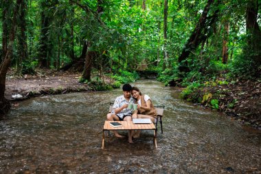 Rahatla ve romantik zaman kavramı, genç Asyalı çift tatilde tropikal ormandaki bir derenin ortasında birlikte oturup kahve içmekten zevk alıyorlar..
