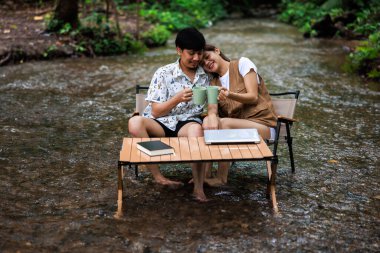 Rahatla ve romantik zaman kavramı, genç Asyalı çift tatilde tropikal ormandaki bir derenin ortasında birlikte oturup kahve içmekten zevk alıyorlar..