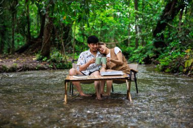 Rahatla ve romantik zaman kavramı, genç Asyalı çift tatilde tropikal ormandaki bir derenin ortasında birlikte oturup kahve içmekten zevk alıyorlar..