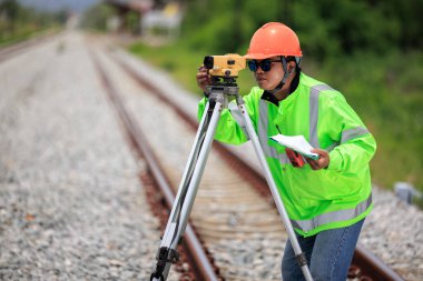 Yeşil üniformalı ve turuncu başlıklı Asyalı mühendis teodolit ekipman ve demiryolu şantiyesinde çalışan inşaat işçilerini gösteren veri kağıdı kullanıyor. Onarım ve bakım demiryolu hatları. 