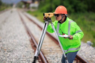 Yeşil üniformalı ve turuncu başlıklı Asyalı mühendis teodolit ekipman ve demiryolu şantiyesinde çalışan inşaat işçilerini gösteren veri kağıdı kullanıyor. Onarım ve bakım demiryolu hatları. 