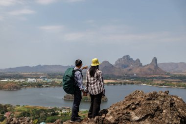 Kayalık bir zirvede el ele tutuşan gezgin bir çiftin arka planda bir rezervuar, bir kasaba ve dağlar barındıran manzaraya bakışları. Bu paylaşılan bir macera ve açık hava romantizmi hissi veriyor..
