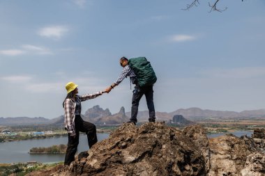 Kayalık bir zirvede el ele tutuşan gezgin bir çiftin arka planda bir rezervuar, bir kasaba ve dağlar barındıran manzaraya bakışları. Bu paylaşılan bir macera ve açık hava romantizmi hissi veriyor..