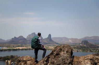 Kayalık bir zirvede duran erkek bir gezginin geniş bir görüntüsü. Sırt çantası takmış ve aşağıdaki manzaraya bakıyor. Bu manzarada bir rezervuar, bir kasaba ve arka planda dağlar var. Bu uçsuz bucaksız doğada bir keşif ve macera hissi taşır..