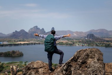 Kayalık bir tepenin üzerinde duran, sırt çantası takan ve kollarını açan, belki de rüzgarı ya da manzarayı kucaklayan bir erkek gezginin geniş bir görüntüsü. Arka planda bir rezervuar, bir kasaba ve dağlardan oluşan özgürlük ve macera duygusu yer alıyor..