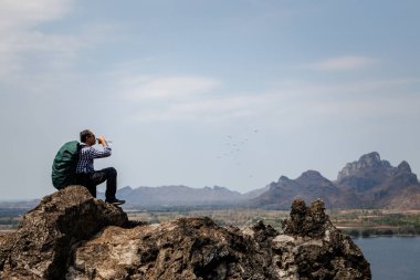 Bir erkek gezginin sırt çantası, içme suyu ile kayalık bir zirvede dinlendiği ve benzersiz dağlar ve mavi bir gökyüzünün yer aldığı manzaraya baktığı görülüyor. Bu görüntü, doğadaki bir doğa macerası sırasında bir dinlenme duygusu taşır..