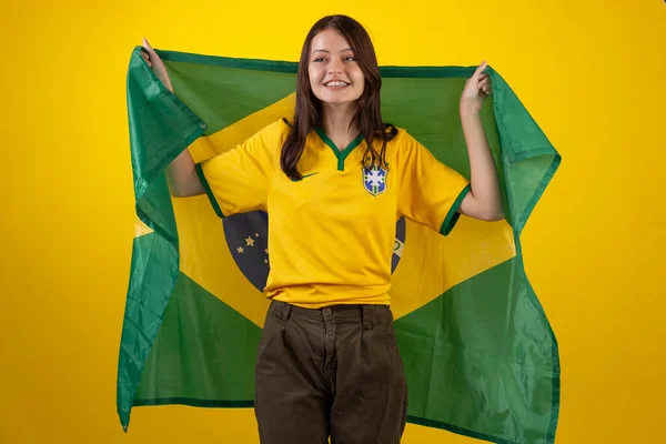 oung woman wearing the official uniform shirt of the Brazilian soccer team in the 2022 Qatar Cup and with the flag of Brazil in studio photo. Brazilian fan.