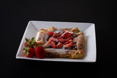 Çilekli börek, çilekli pizza hamurlu tatlı fındıklı krema gibi çikolatalı, beyaz bir tabağın içindeki siyah kumaşın üstünde.