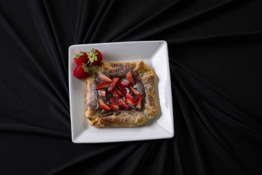 Çilekli börek, çilekli pizza hamurlu tatlı fındıklı krema gibi çikolatalı, beyaz bir tabağın içindeki siyah kumaşın üstünde.