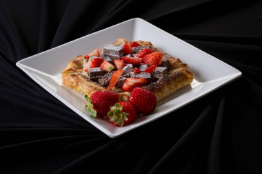 Çilekli börek, çilekli pizza hamurlu tatlı fındıklı krema gibi çikolatalı, beyaz bir tabağın içindeki siyah kumaşın üstünde.