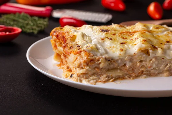 slice of lasagna with melted cheese on top and chicken meat filling, on a black table in a white plate, surrounded by kitchen utensils, tomatoes and red pepper