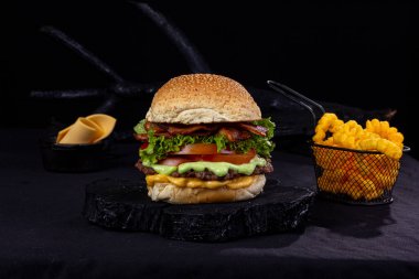 Büyük hamburger, içinde patates kızartması olan küçük demir bir sepet, siyah kumaşlı ve ahşap kumaşlı bir masanın üzerinde desen ve siyah renkli,