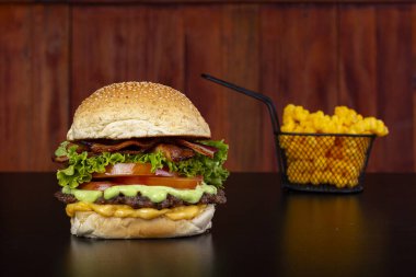 Yanlarında siyah bir masada, yansıması ve ahşap arka planı olan patates kızartması içeren demir bir sepetle dev bir hamburger.