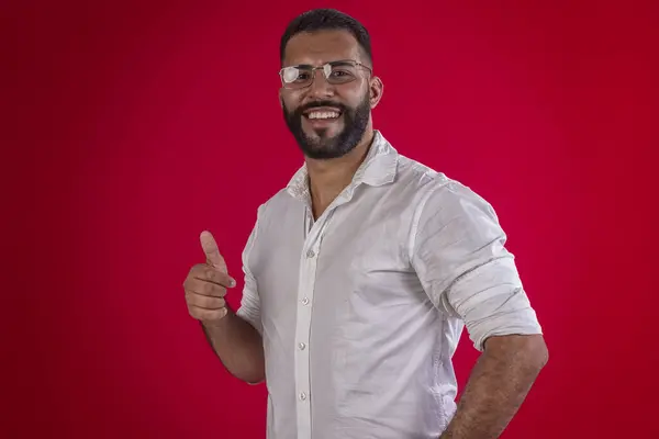 Adamım, sakallı, gözlüklü, beyaz gömlekli, değişik yüz ifadeli, kırmızı stüdyo arka planda.