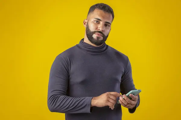 Sakallı, siyah tişörtlü, elinde cep telefonu olan, farklı yüz ifadeleri olan bir adam.