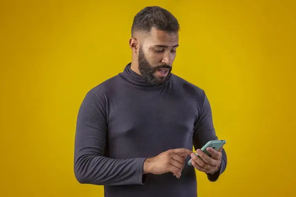 Sakallı, siyah tişörtlü, elinde cep telefonu olan, farklı yüz ifadeleri olan bir adam.