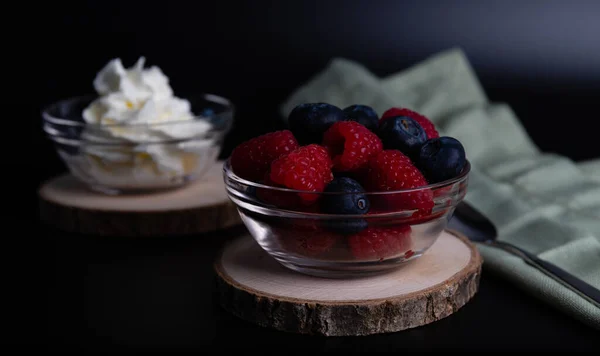 Blueberries and raspberries in a glass bowl also a glass bowl with whipped cream on a dark background with textile. Macro shooting, all focus on berries.