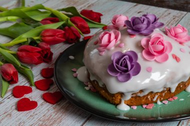 Heart-shaped cake with lemon icing decorated with pink and purple flowers with fresh red tulips on the table