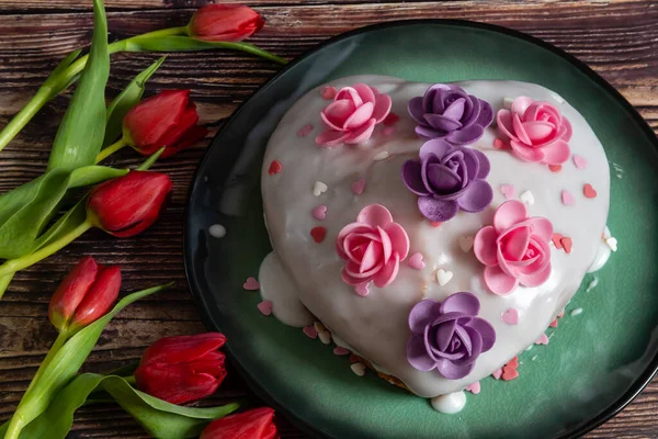 Heart-shaped cake with lemon icing decorated with pink and purple flowers with fresh red tulips on the table