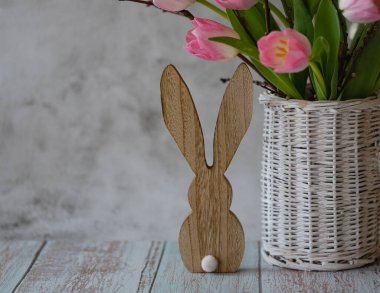 Twigs and pink tulips in a vase of willow branches and a wooden bunny on a light background, Easter greeting card, copy space.