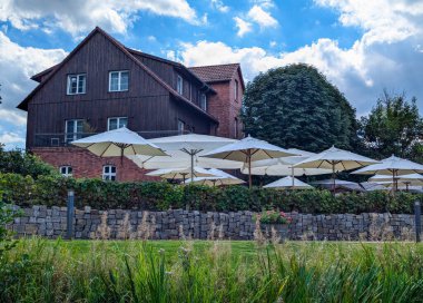 Almanya, Brandenburg, Schlepzig, 16 Ağustos 2024: Nehir kenarındaki yemyeşil manzaraya tepeden bakan çok sayıda şemsiyeli bir açık hava yemek alanı, mobil fotoğraf.