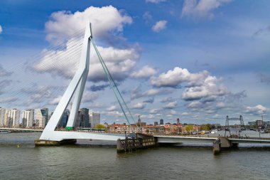 Rotterdam, Hollanda - 1 Nisan 2024: Erasmus Köprüsü, arka planda Rotterdam silueti ve açık, parçalı bulutlu bir günde aşağıda seyahat eden teknelerle Maas Nehri boyunca uzanır..