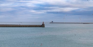 Zeebrugge, Belçika-2 Nisan 2024, denizin üzerinde uçan martılarla dolu bir dalgakıran üzerinde kırmızı ve beyaz bir deniz feneri duruyor. Rüzgar türbinleri bulutlu bir gökyüzünün altında dönüş yapar.