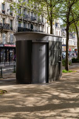 Paris, Fransa-14 Nisan 2025: Gri bir kendi kendini temizleyen umumi tuvalet gölgeli park alanındaki apartman binaları ve yakınlarındaki dükkanlarda duruyor.