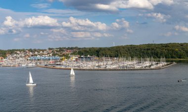 Kiel, Almanya-09.08.2025: Büyük marina, kıyı kasabası evleri ve bulutlu gökyüzünün altındaki yeşil ormanlık tepelerle çevrili yelkenli ve yatlarla dolu.