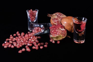 Bardakta kırmızı nar suyu, nar meyvesi dilimleri ve siyah arkaplana saçılmış tohumlar..