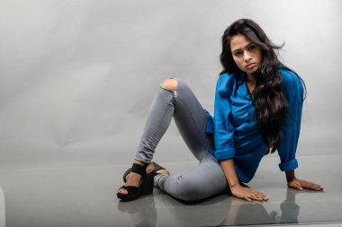 Gri arka plan karşısında gündelik giyimli Hintli genç manken. Stok fotoğrafı. Uzun siyah saç modeli mavi gömlek, gri kot pantolon ve siyah topuklu ayakkabı giyiyor..