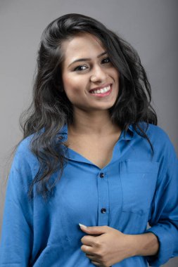 Gri arka plan karşısında gündelik giyimli Hintli genç manken. Stok fotoğrafı. Uzun siyah saç modeli mavi gömlek giyiyor..