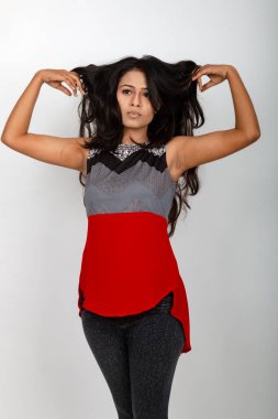 Genç, güzel bir Hintli kadın modelin gündelik kıyafetleriyle stüdyo fotoğrafı. Kırmızı ve gri tasarımcı bluzu ve beyaz arka planda siyah kot pantolon giyiyor. Kadın manken. Moda Portresi.