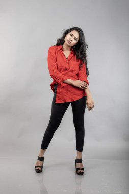 Gri arka plan karşısında gündelik giyimli Hintli genç bayan model - stok fotoğrafı. Kırmızı gömlek ve siyah kot giyen uzun siyah saçlı bir manken..