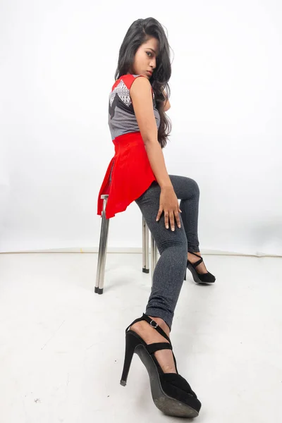 Studio shot of a young, beautiful Indian female model in casual wear wearing red and grey designer top and black jeans against white background. Female model. Fashion Portrait.